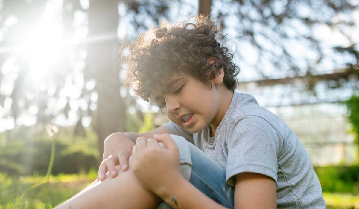 Upset kid seated on the grass in the park touching his injured knee-cap with both hands